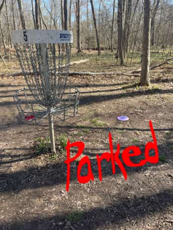 Disc golf disc on the ground about 10 feet from a disc golf backet - hole 5
